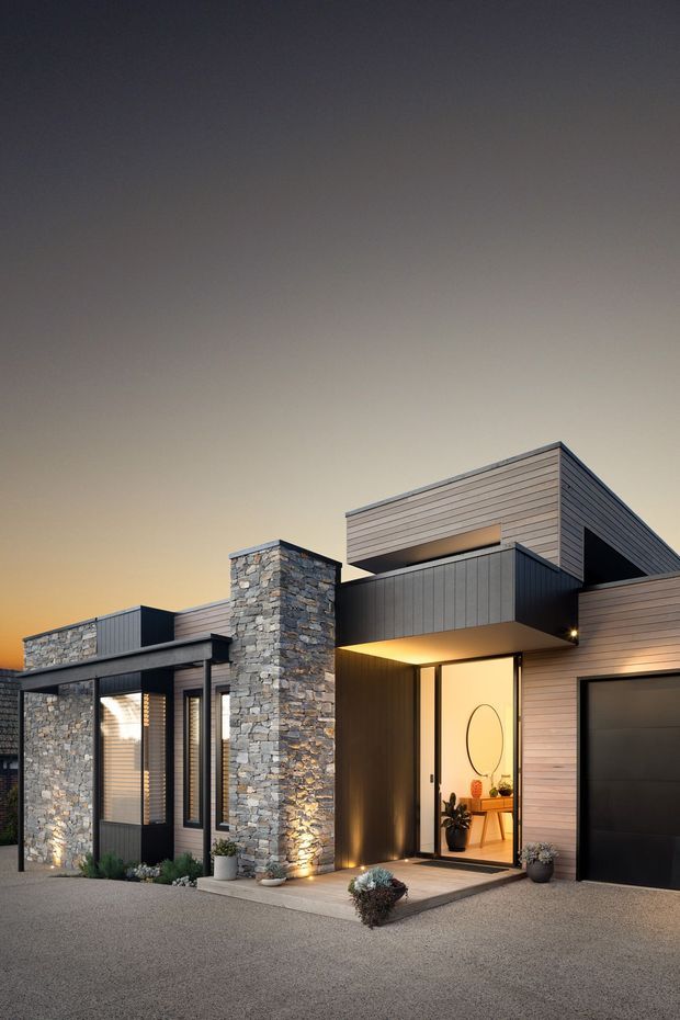 Contemporary single-storey residential facade photographed at dusk, featuring dark vertical and horizontal cladding, extensive natural stacked-stone masonry, a projecting upper volume, recessed entry with warm lighting, narrow windows, and an integrated dark garage door. Minimal landscaping and a paved forecourt complete the modern exterior composition.