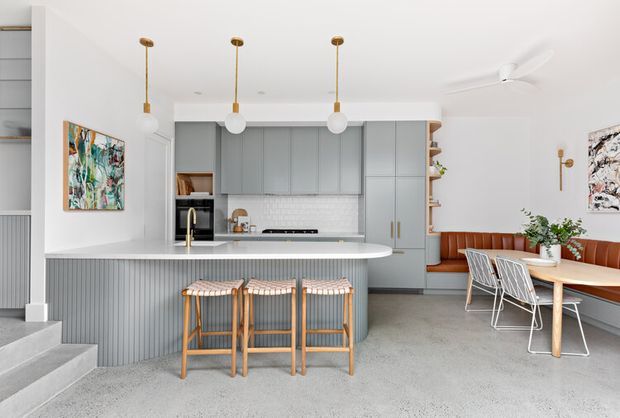 Bright open-plan kitchen and dining area with a curved island clad in vertical grey paneling, three timber stools, pale grey built-in cabinetry, white brick backsplash, integrated appliances, brass pendant lights, and a built-in tan leather dining nook. Polished concrete flooring, white walls, colourful artwork, and contemporary furnishings create a clean, refined space.