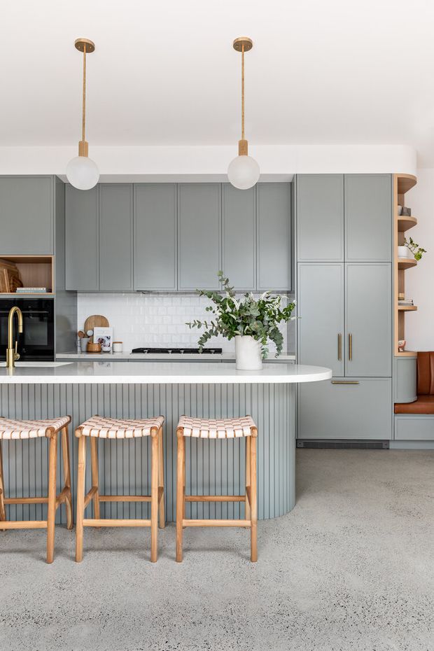 A bright, refined kitchen with muted grey-green full-height cabinetry, a white tiled splashback, a pale stone island with vertically fluted panel detailing, and integrated appliances. Three timber-framed woven bar stools sit at the island. Brass pendant lights with white globe shades hang above, while open curved shelving and a built-in seating nook add warmth on the right. The floor appears to be a light speckled concrete or terrazzo-like finish.