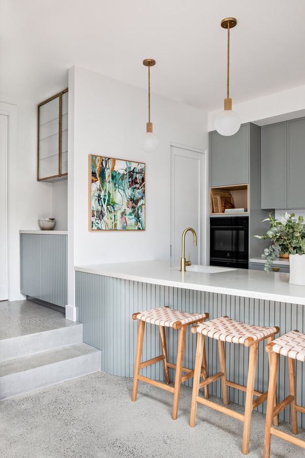 Bright contemporary kitchen with a white stone island benchtop, muted grey-green vertically panelled island cabinetry, integrated grey cabinetry, brass gooseneck tapware, pendant globe lights, and three woven timber bar stools. The space connects to a raised hallway via polished concrete steps and includes a colourful abstract artwork and indoor greenery.