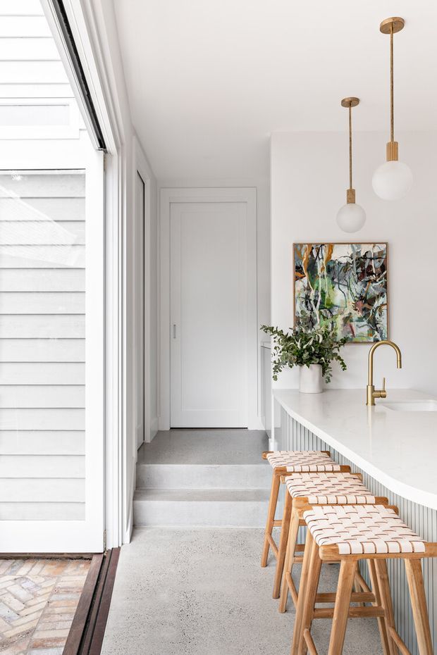 Bright, refined kitchen and entryway interior with a long white island, integrated ribbed cabinetry, brass gooseneck tap, and woven-seat timber stools. A large glazed opening connects to a covered exterior walkway, while white wall panelling, pendant lights, and an abstract artwork add detail. The floor is a smooth grey concrete or terrazzo-like finish, with a tiled threshold at the opening.