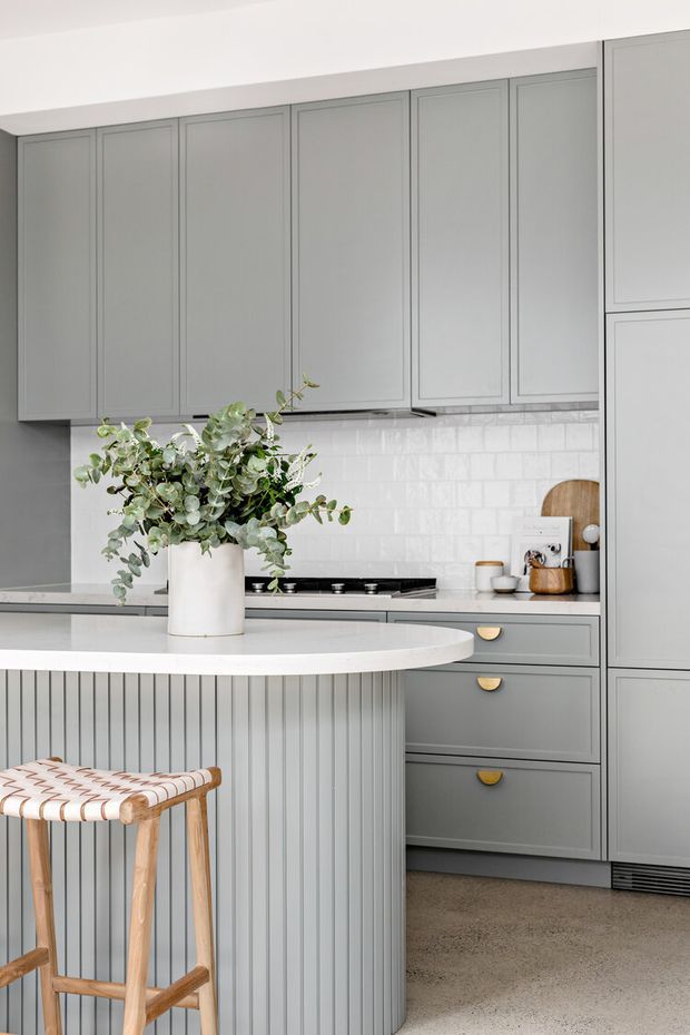 A refined contemporary kitchen with full-height grey cabinetry, recessed-panel doors, brass cup pulls, a white tiled backsplash, and a pale curved island with vertically fluted detailing. A timber bar stool and a large eucalyptus arrangement add natural warmth to the otherwise soft neutral palette.
