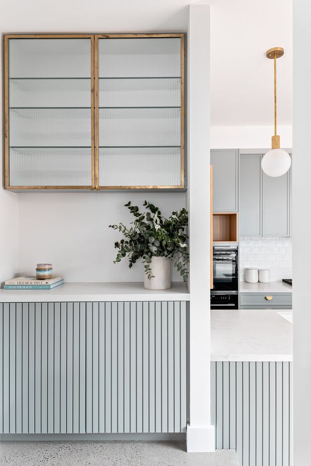 Bright, refined kitchen interior featuring pale grey vertically fluted cabinetry, a white stone benchtop, white tiled splashback, and a timber-framed glass-front display cabinet. A sculptural brass-and-white pendant hangs above the kitchen, while a built-in ledge displays books and a leafy plant. The space has a clean contemporary look with subtle traditional detailing and warm timber accents.