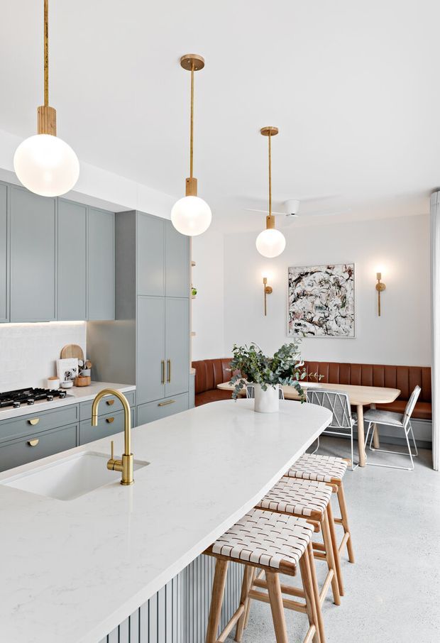 Bright, refined open-plan kitchen and dining area with muted blue-grey shaker cabinetry, a large white stone island with integrated sink and brass tapware, woven timber bar stools, and a built-in brown leather banquette. Globe pendant lights hang above the island, while an abstract artwork and wall sconces add visual interest to the dining nook.