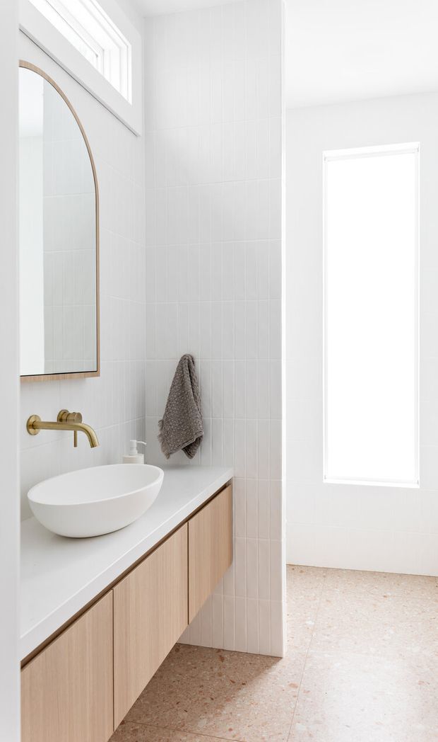 Bright, minimalist bathroom featuring a floating light timber vanity with a white vessel basin, wall-mounted brass tapware, white tiled walls, a rounded timber-framed mirror, and terrazzo-style flooring. A tall frosted or overexposed window provides abundant natural light.