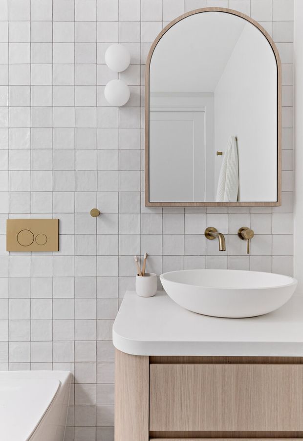 A refined contemporary bathroom with a white grid-tile wall, arched timber-framed mirror, floating light-wood vanity, white vessel basin, and wall-mounted brushed brass tapware. A concealed cistern flush plate and partial toilet are visible at left, while the mirror reflects a white door and towel.
