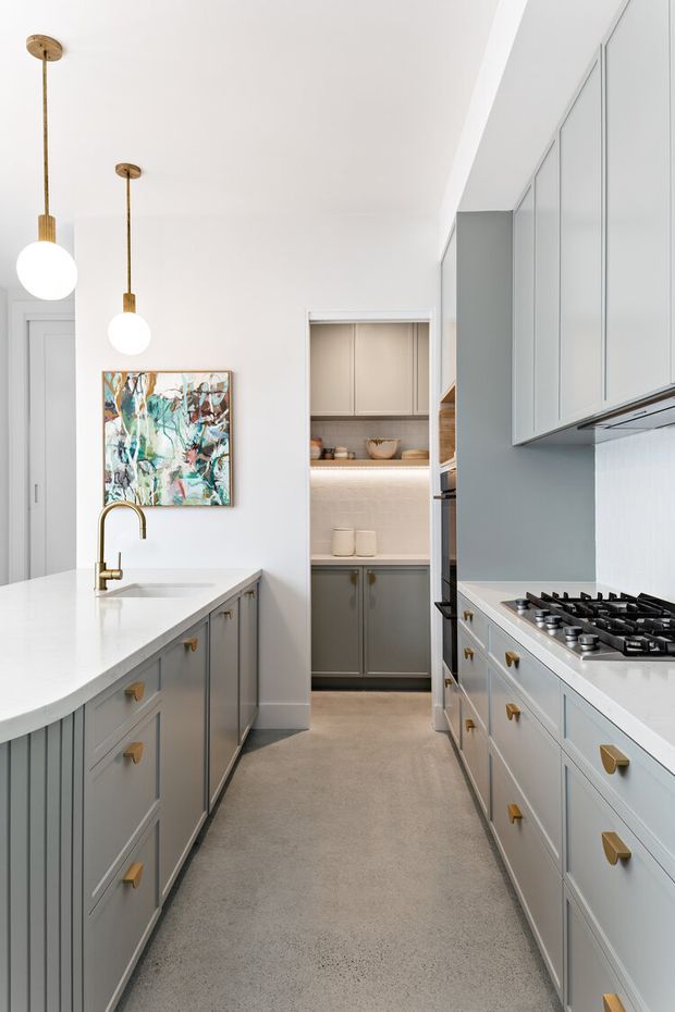 A bright galley-style kitchen with extensive muted grey built-in cabinetry, white stone benchtops, integrated appliances, and a large gas cooktop. Brass or gold-toned pendant lights, tapware, and cabinet pulls add warmth. A compact walk-through pantry or scullery is visible at the rear, with open shelving and under-cabinet lighting. The kitchen has white walls, a light neutral floor, and a colourful abstract artwork.
