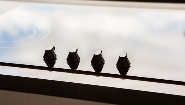 Close-up of a sleek black glass cooktop with four black conical control knobs arranged in a row. The reflective surface and minimal detailing suggest a modern built-in cooking appliance.