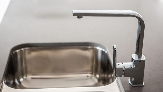 Close-up product-style image of a contemporary kitchen sink and single-lever gooseneck faucet. The sink and faucet are stainless or chrome-finished metal, set against a neutral grey benchtop and light background.