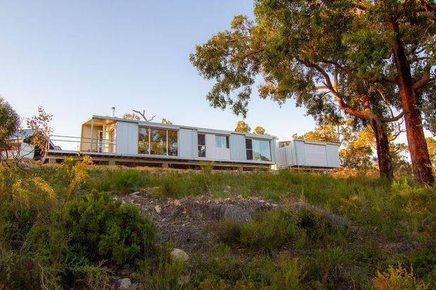 A long, low-slung contemporary modular-style house set on a rocky, vegetated sloping site. The building has pale light-coloured cladding, extensive floor-to-ceiling glazing, flat roof sections, and appears elevated on a narrow structural platform. Large mature trees and native landscaping surround the residence.