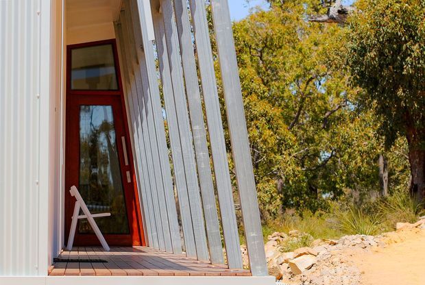 Exterior view of a contemporary building entrance with a tall red-framed glazed door, timber deck, and a series of vertical corrugated metal fins or screens along the facade. The structure sits beside a dry, rocky native landscape with mature trees and grasses.
