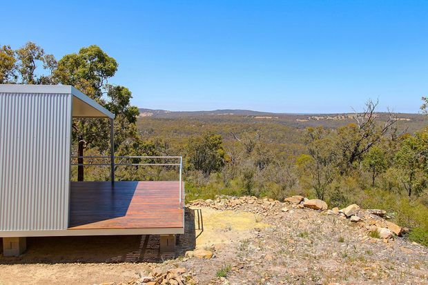Exterior view of a compact modern cabin or pavilion with corrugated metal cladding and a timber deck projecting toward a broad bushland landscape. The structure has a simple low-pitched roof, open-sided covered deck, dark metal balustrade, and elevated construction on supports. Dry rocky terrain and native woodland surround the building under a clear blue sky.