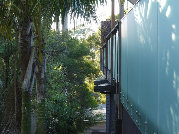 Exterior side view of a contemporary building nestled among dense tropical vegetation. The facade combines large pale blue-green flat panels with dark vertical cladding, tall glazing, and projecting balcony or deck elements. Palm-like trees and lush greenery closely frame the structure, creating a secluded, elevated site impression.