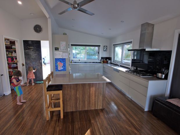 A bright, open-plan residential kitchen viewed from the living area. The room features a large central island with a pale stone benchtop and timber-look cabinetry, extensive white built-in cabinetry, a black tiled splashback, stainless-steel rangehood, gas cooktop, multiple windows, and dark timber flooring. A pantry and adjacent living area are visible beyond the kitchen.