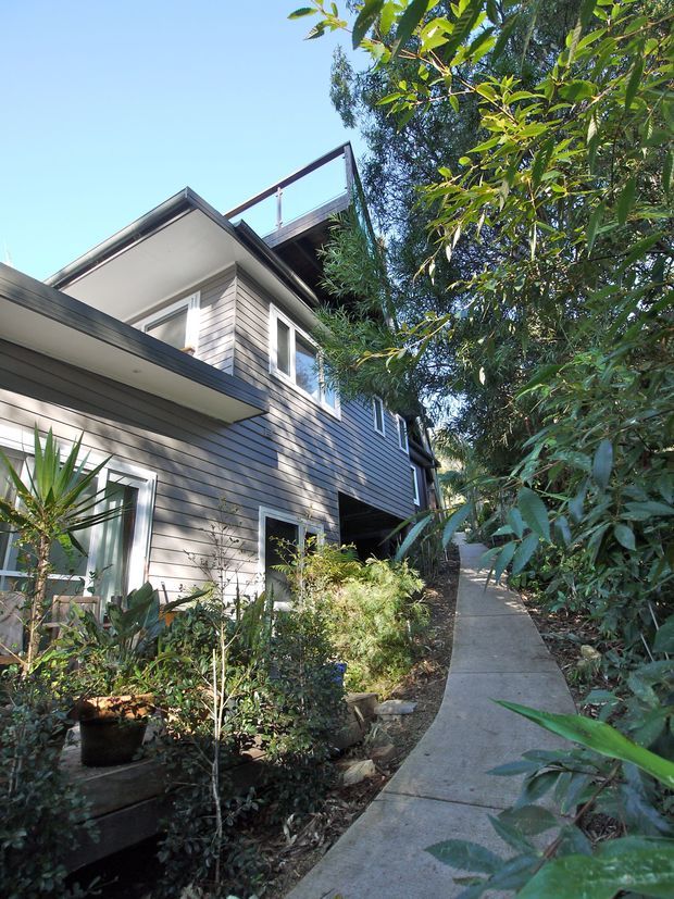 Exterior view of a multi-level contemporary house set within dense garden landscaping. The building has horizontal grey weatherboard cladding, white-trimmed windows, projecting flat-roofed sections, and an upper terrace or balcony with a transparent balustrade. A narrow concrete walkway leads alongside the planted facade.