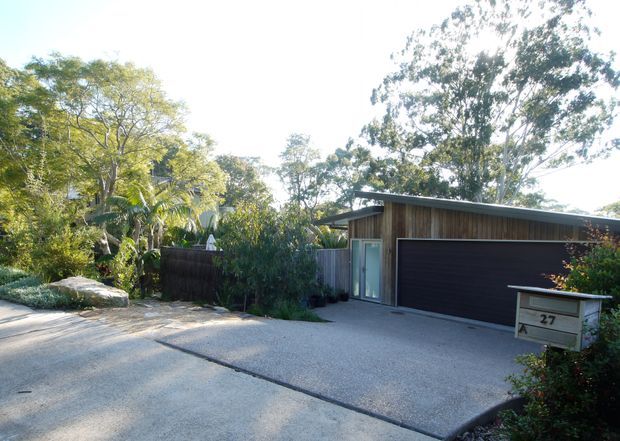 Street-level exterior view of a low-profile contemporary residence, dominated by a broad dark garage door, vertical timber-clad facade, and a simple shallow-sloping roof. A glazed entry door sits beside the garage, with a concrete driveway, landscaped planting, screening fence, and mailbox in the foreground.