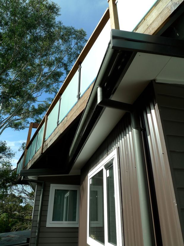 Upward-looking exterior detail of a modern multi-level building, showing a projecting upper balcony or roof edge with timber framing and translucent balustrade panels. The lower facade combines dark horizontal cladding, vertical corrugated metal cladding, white-framed glazing, exposed gutters and dark metal eaves. Mature trees surround the structure.