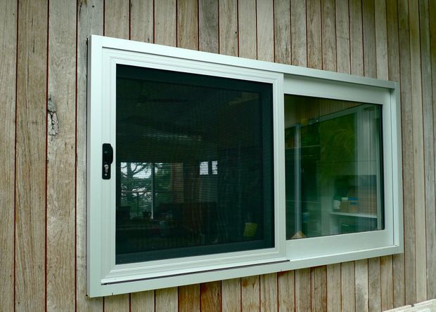 Close-up exterior view of a white-framed sliding window set into vertically clad weatherboard walling. The window has dark insect screening or tinted glazing, visible reflective interior furnishings, and a simple horizontal two-panel configuration. The surrounding timber cladding is weathered and rustic.