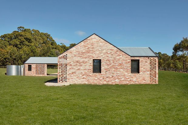 Exterior view of a simple single-storey brick building set in a broad grassy landscape. The main gabled volume has pale red and cream exposed brick walls, dark-framed rectangular windows, and a corrugated metal pitched roof. A lower connected wing and cylindrical metal tank are visible to the side, with mature trees behind.