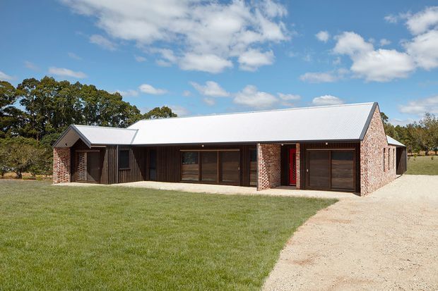 Single-storey rural residence with a long low-pitched corrugated metal gable roof, dark vertical timber-clad sections, and exposed red brick walls. The facade includes large dark-framed glazed openings and an integrated garage door, set beside a broad lawn and gravel driveway in an open countryside setting.