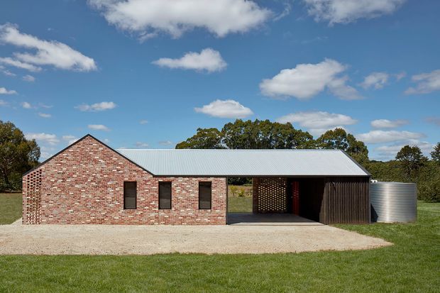 A single-storey rural building with a red and cream brick gabled wing, a long pale corrugated metal roof extending over an open covered area, dark vertical timber cladding, and a cylindrical corrugated metal water tank. The building sits in a grassy landscape under a bright blue sky.