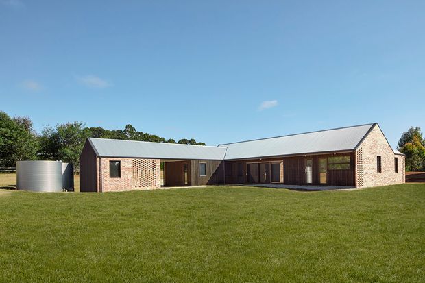 Single-storey rural-looking contemporary building viewed from across a broad lawn. The long low structure has a folded gable roof with pale metal roofing, exposed red brick end walls and sections, dark timber-clad recessed central elevations, and a covered or glazed central courtyard-like frontage. A cylindrical water tank stands to the left, with mature trees behind the property under a clear blue sky.
