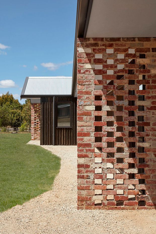 Exterior architectural detail of a contemporary building, featuring a perforated red brick screen wall with alternating open voids and pale infill bricks. Behind it is a dark vertical timber-clad volume with a corrugated metal pitched roof and window. The building sits beside a gravel path and lawn under a clear blue sky.
