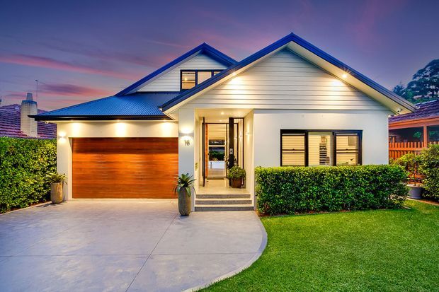 A well-presented suburban house facade photographed at dusk, featuring white horizontal weatherboard cladding, a blue corrugated pitched roof with intersecting gables, warm exterior lighting, a timber garage door, and a sheltered front entry. A curved concrete driveway and neatly trimmed hedges frame the single-storey home.