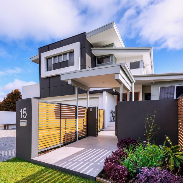 Contemporary multi-storey residential facade with crisp white and dark charcoal cladding, projecting flat-roofed volumes, deep covered entry, and horizontal timber-look slatted fencing and gates. A paved path leads through a landscaped front garden to the entry, with the house number displayed on a dark rendered pillar.