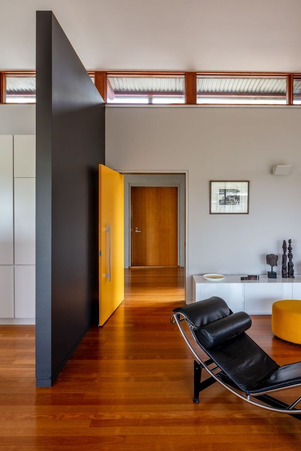 Interior view of a refined mid-century-inspired living room or entry area with warm timber flooring, a dramatic dark partition wall, clerestory windows, timber doors, a bright yellow entrance door, and a black leather sling lounge chair. A white low sideboard and yellow ottoman add modern colour accents.