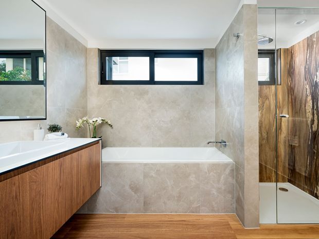 Contemporary bathroom featuring a freestanding-style bathtub beneath a high horizontal window, a timber-look floating vanity, beige stone-look wall tiling, and a glass-enclosed shower with dramatic dark timber-look wall finish. A large mirror reflects the vanity and window, while warm timber flooring adds contrast.