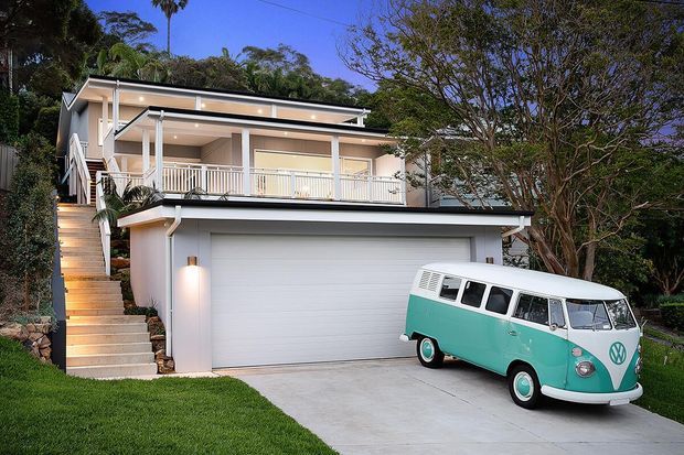 A two-storey residential house on a sloping, landscaped site, with a large white garage at ground level, an external stairway, and an elevated upper-level veranda with white railings and slim posts. The home has a light contemporary facade, broad roof overhangs, and recessed exterior lighting. A vintage turquoise-and-white van is parked in the driveway, with mature trees and lawn surrounding the property.