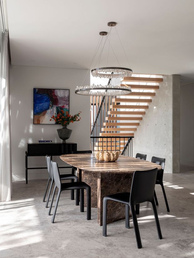 A refined open-plan dining area with a large oval dark marble dining table surrounded by black dining chairs. Two suspended circular crystal chandeliers hang above the table. The room features pale plaster walls, a polished light concrete or stone floor, expansive sheer-curtained glazing, an abstract artwork, and a black sideboard. An open floating timber stair with black metal balustrading forms a strong architectural feature in the background.