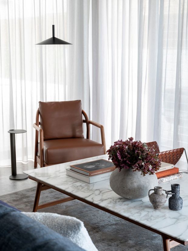 A softly lit, refined living room vignette with sheer floor-to-ceiling curtains, a brown leather lounge chair, a marble-topped timber coffee table, sculptural ceramic vessels, books, and a dark pendant light. The palette is warm neutral with muted brown, grey, white, and black accents, creating a calm contemporary atmosphere.