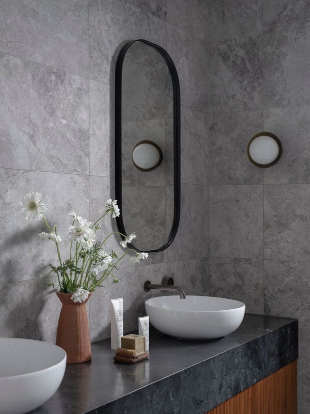 A refined bathroom vanity featuring a dark stone benchtop, white vessel basins, wall-mounted dark metal tapware, and a tall arched black-framed mirror. Large grey stone-look wall tiles create a moody, luxurious backdrop, with warm timber cabinetry below and subtle wall sconces. A ceramic vase with flowers and toiletries add styling details.