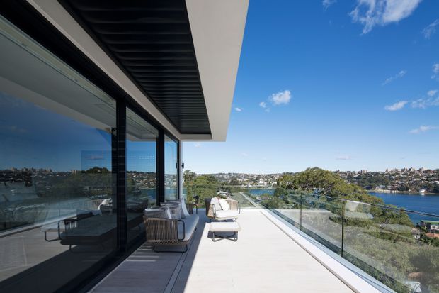 A contemporary waterfront terrace or balcony viewed from outside a modern residence. The broad pale terrace has glass balustrades, outdoor lounge seating and sun loungers, while full-height dark-framed glazing reflects the seating and opens onto the interior. A strong projecting roof slab with a dark ribbed soffit provides shade, and the terrace overlooks a river or harbour and densely built opposite shoreline.