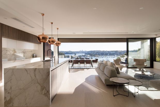 A bright, open-plan contemporary interior combining a kitchen, dining area and living room, with expansive glazing overlooking a waterfront landscape. The kitchen features a large marble-clad island, light wood cabinetry, integrated appliances and copper pendant lights. A neutral-toned sofa and lounge seating occupy the living area, while sliding glass doors open toward an outdoor terrace or balcony.