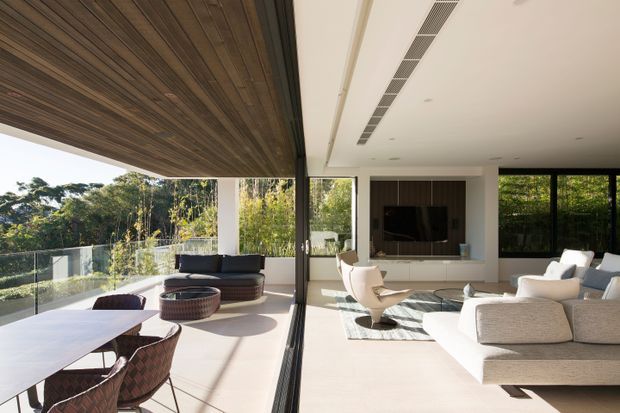 A bright contemporary open-plan living and dining area opening onto a covered terrace through expansive floor-to-ceiling glazing. The interior features a large light upholstered sectional sofa, sculptural lounge chairs, a wall-mounted television with built-in cabinetry, and a dining table with woven chairs. A timber-lined soffit extends over the terrace, contrasting with the white ceiling and pale flooring, while lush greenery is visible beyond the glass balustrade.