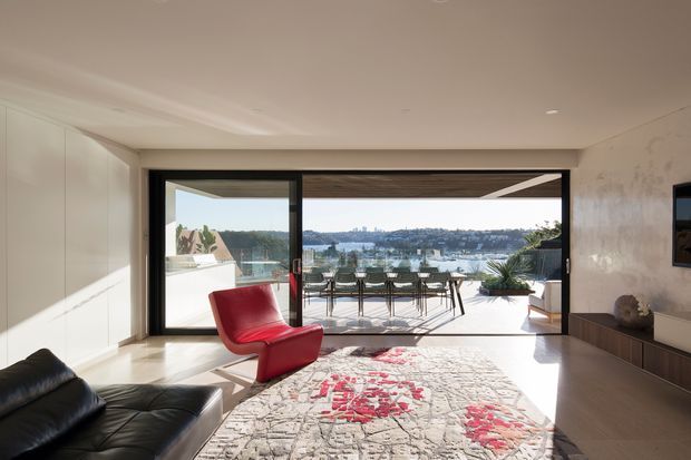 Bright contemporary living room opening through large black-framed sliding glass doors to a waterfront-facing terrace. The room has pale walls and cabinetry, warm timber flooring, a black leather sofa, a sculptural red lounge chair, a large patterned rug, and low built-in media cabinetry. Outdoor dining furniture, planting, water, and a distant urban shoreline are visible beyond the glazing.