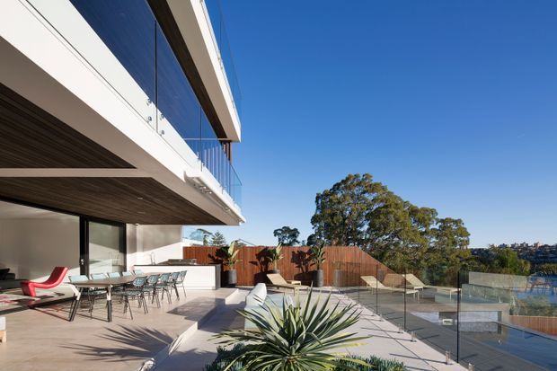 A spacious contemporary multi-level terrace overlooking a landscaped setting and water beyond. The terrace features large-format pale paving, frameless glass balustrades, outdoor dining furniture, sun loungers, built-in outdoor cabinetry, timber soffit detailing, and extensive glazing connecting to the interior. A projecting upper level with clear glass railing creates a strong horizontal architectural form.
