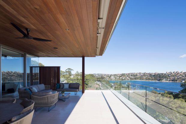 A spacious covered outdoor terrace overlooking a blue-water bay, furnished with low lounge seating and woven armchairs. The terrace has a warm timber-lined soffit with an integrated ceiling fan, slender dark posts, expansive glazing, and a frameless glass balustrade. The contemporary home appears elevated above a coastal urban setting, with strong indoor-outdoor connection and clear views across the water.