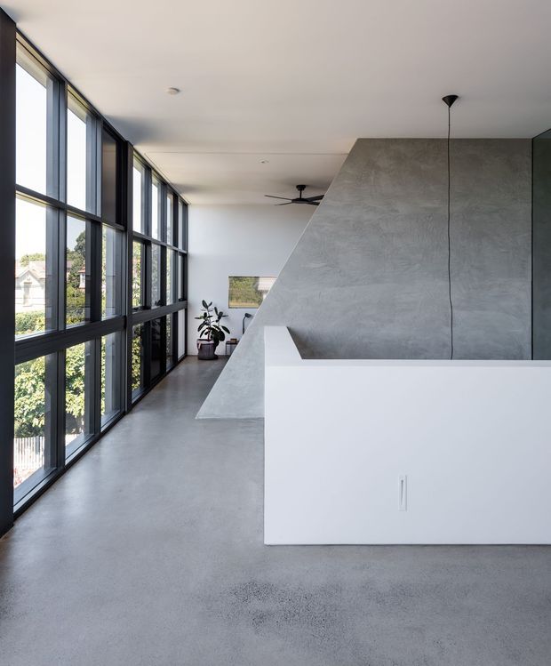 A bright, minimalist open-plan interior with a long wall of black-framed floor-to-ceiling glazing on the left. A sculptural angular partition or stair enclosure in textured grey concrete-like finish divides the space, with a white built-in island or joinery volume in the foreground. The room has a smooth grey concrete floor, white ceiling, sparse furnishings, and a contemporary architectural character.