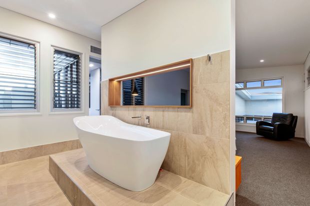 A bright contemporary bathroom featuring a freestanding white bathtub set on a raised tiled platform, pale beige stone-look floor and wall tiles, and a timber-framed recessed mirror above the bath. Timber venetian blinds cover the windows, and an adjoining carpeted room with a lounge chair is visible through the opening.