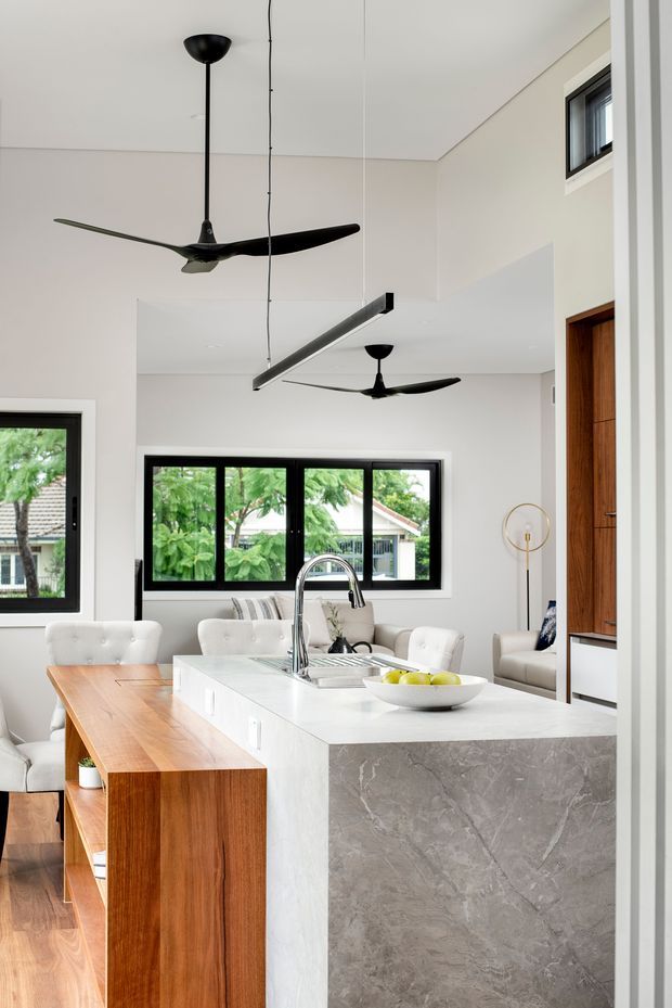 Bright open-plan kitchen and living area viewed from the kitchen, featuring a large white stone island with waterfall-style grey-veined sides, integrated sink and curved chrome tap, warm timber cabinetry and dining bench, black-framed windows, pale walls, and two black ceiling fans. A neutral upholstered seating area is visible beyond the island.