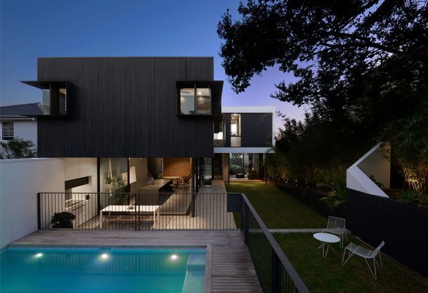 Twilight exterior view of a contemporary multi-level residence, featuring a dark vertically ribbed upper volume, extensive glazing, a recessed lower outdoor dining area, timber pool deck, illuminated swimming pool, landscaped lawn, and a sculptural white-and-dark garden structure. The composition highlights the house facade, outdoor living spaces, and strong indoor-outdoor connection.