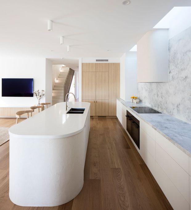 Bright contemporary open-plan kitchen with a large sculptural white island, integrated sink and tap, pale timber cabinetry, white cabinetry, marble-look stone benchtops, and a textured light stone feature wall. Timber flooring continues into the adjacent dining and living area, with stairs and a wall-mounted television visible beyond.