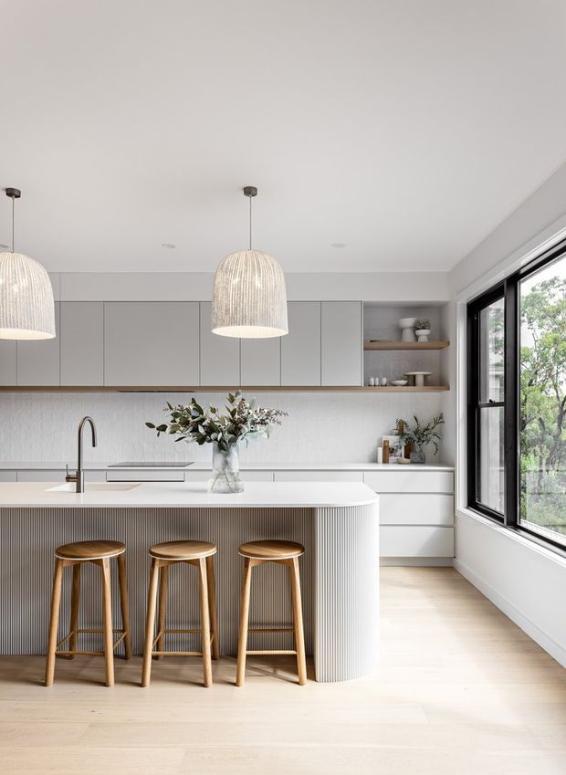 Bright contemporary kitchen with a large island featuring a vertically fluted light-grey base, pale stone benchtop, integrated sink and tap, and three timber bar stools. Full-height light-grey cabinetry, open timber shelving, a subtly textured backsplash, pendant lights, and light timber flooring create a clean, softly neutral interior. Black-framed glazing on the right provides abundant natural light and views to greenery.