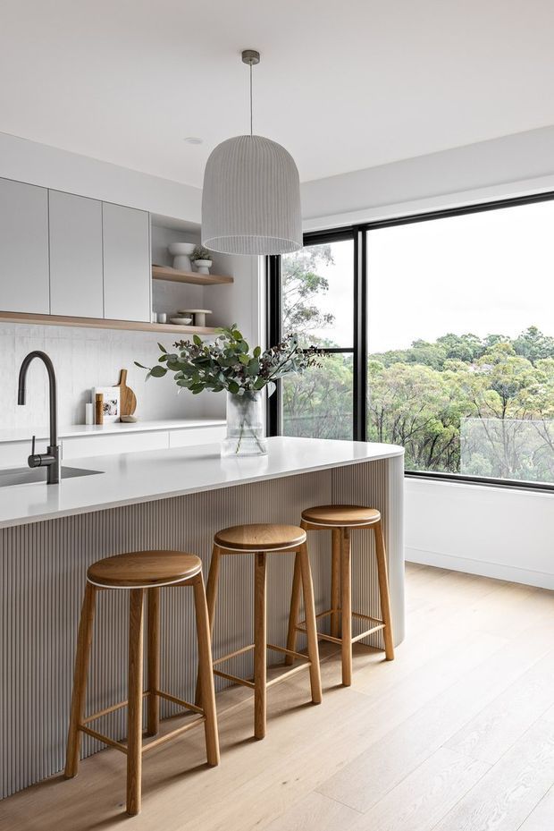 Bright contemporary kitchen with a large white island, integrated sink and curved black tap, vertically fluted island cabinetry, pale timber flooring, light grey overhead cabinetry, open timber shelving, and a textured pendant light. Three timber bar stools line the island, while expansive black-framed glazing overlooks leafy trees.
