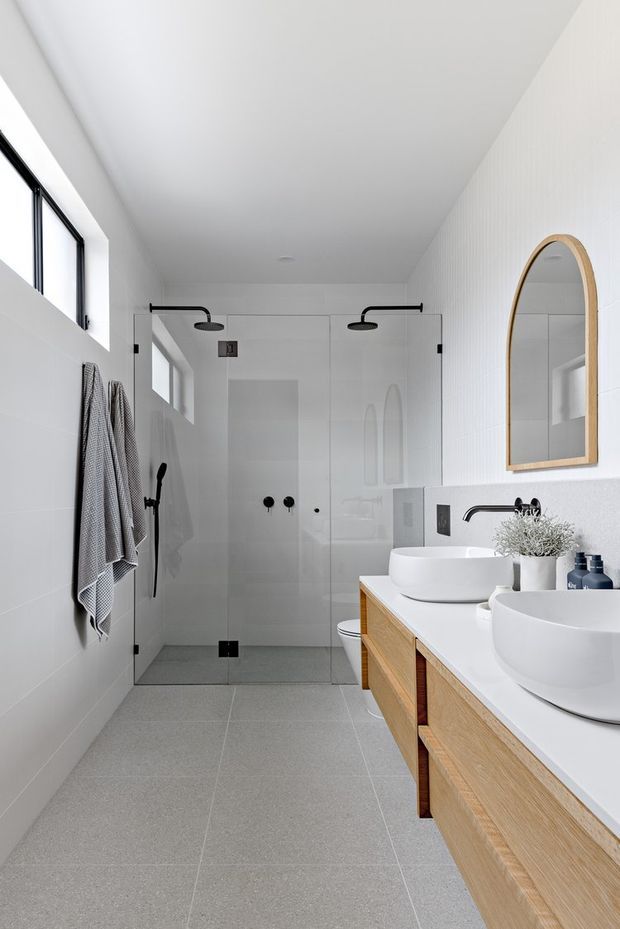 Bright contemporary bathroom featuring a walk-in glass shower with matte black fixtures, twin white vessel basins on a light timber vanity, large grey floor tiles, white wall surfaces, and an arched timber-framed mirror. Natural light enters through high windows, creating a clean, airy space.