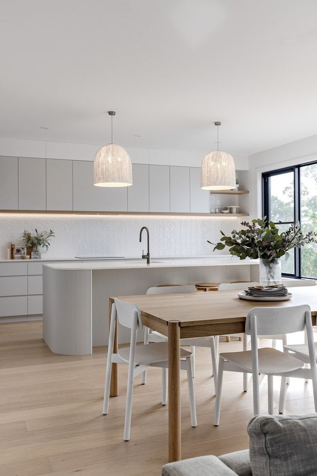 Bright contemporary open-plan kitchen and dining area with pale timber flooring, matte light-grey cabinetry, a white textured tile splashback, and a large curved island clad in vertical ribbed detailing. Two woven pendant lights hang above the island, while a light timber dining table with white chairs sits in the foreground beside large black-framed windows. Neutral, natural materials and abundant daylight create a calm, refined interior.